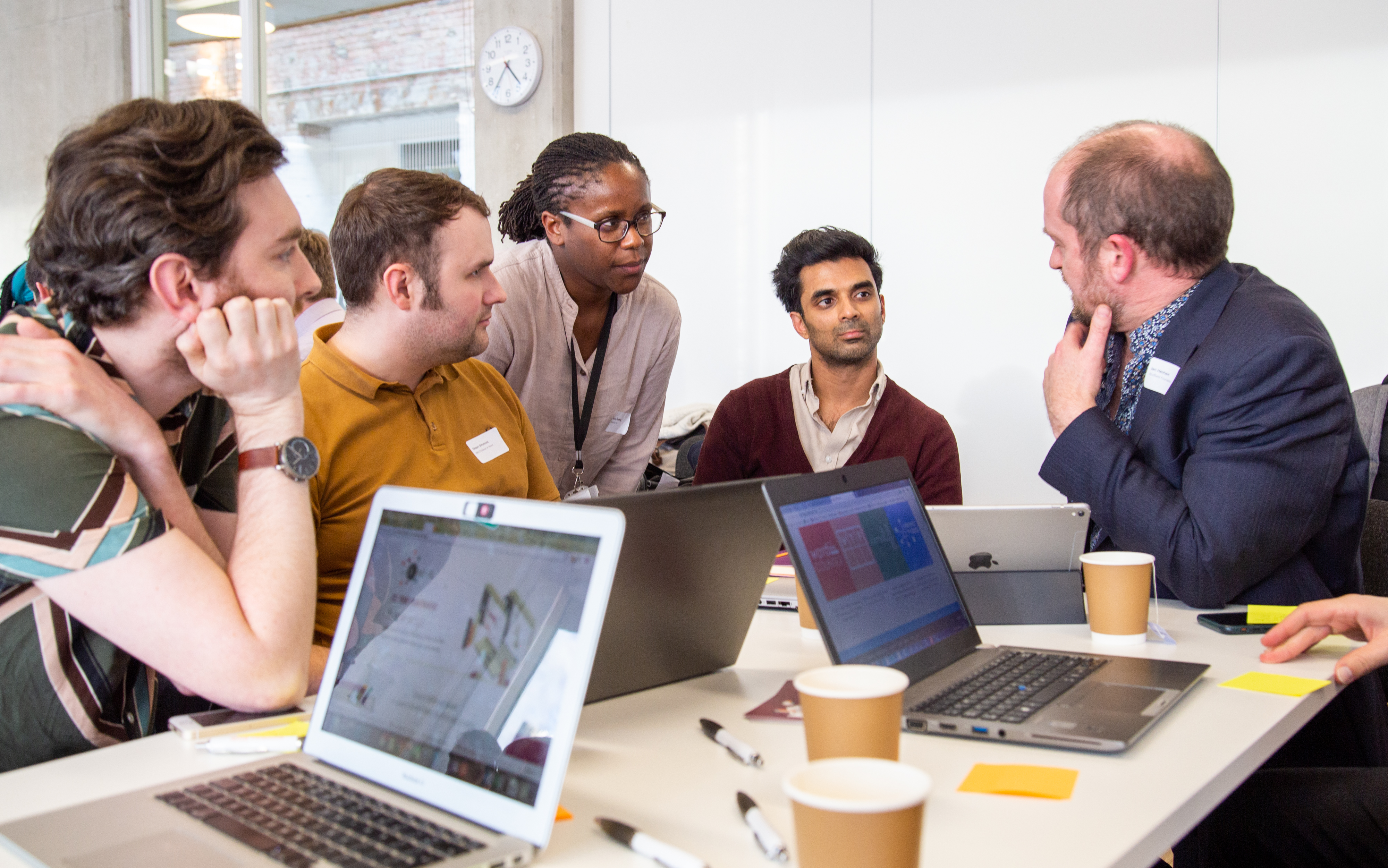 Group of people sat around a desk engaging in discussion with laptops on the table. 